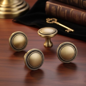 May include: Four antique brass cabinet knobs with a circular design and beaded trim are displayed on a wooden surface. One knob is a handle style. A vintage key and books are in the background. The knobs have a classic, aged appearance.