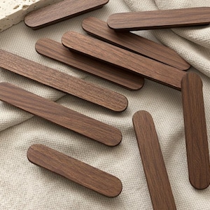 Puede incluir: Una colección de tiradores de armario de madera marrón oscuro con extremos redondeados. Los tiradores están dispuestos sobre una superficie de tela de color neutro. La veta de la madera es visible en cada tirador, mostrando la textura natural y las variaciones de color.