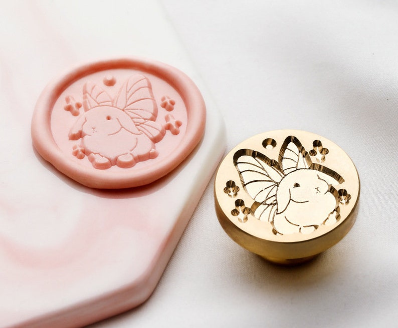 Puede incluir: Un sello de metal dorado con un dise&ntilde;o de un conejo con alas y flores. El sello se utiliza para crear sellos de cera.