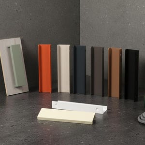 May include: A selection of cabinet pulls in a range of colours: green, orange, cream, navy, brown, and black. Some are mounted on a wooden board. Additional pulls are laid flat. A small potted plant and a rope are also present.