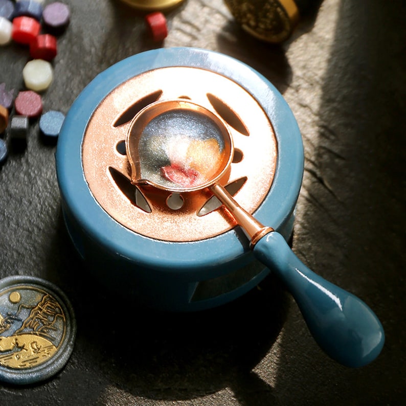 Pu&ograve; includere: Un pentolino per sciogliere la cera in ceramica blu con un bordo color rame e un design decorativo a taglio. Un cucchiaio per sciogliere la cera color rame con un manico in legno blu si trova sopra il pentolino.