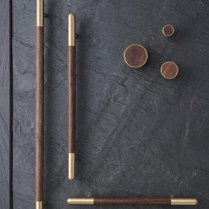 May include: A collection of wooden cabinet hardware with brass accents. The set includes long, slender pulls and round knobs. The wood is a rich brown, and the metal is a shiny gold color. The hardware is arranged on a dark gray surface.