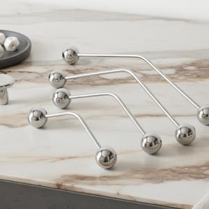 以下が含まれることがあります： 磨かれたシルバーメタルのキャビネットプルとキノコ型のノブのコレクションが、大理石のカウンタートップに配置されています。プルは両端にボールがあり、湾曲したデザインです。小さなプレートには、白い幾何学模様のキャンディーが入っています。
