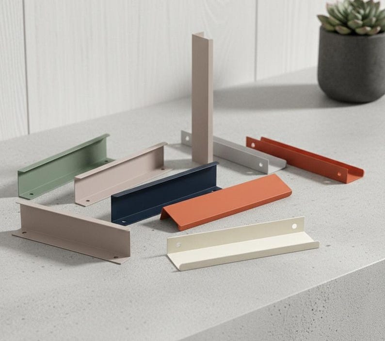 May include: A selection of colourful metal cabinet pulls in shades of green, pink, navy, and orange. The pulls have a modern, minimalist design, with a simple L-shape. Some are upright, while others are lying flat. Each pull has a hole for mounting.