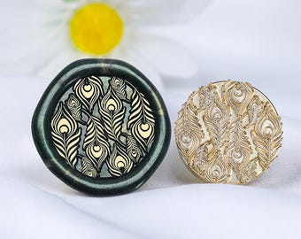 May include: Two wax seal stamps featuring peacock feather designs. One is dark green with cream feathers, the other gold. Both are round and rest on a white surface. The stamps are for decorative use.