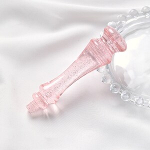 Puede incluir: Un mango de soporte para pastel de vidrio rosa con un acabado brillante. El mango tiene forma de corona y tiene un dise&ntilde;o decorativo.