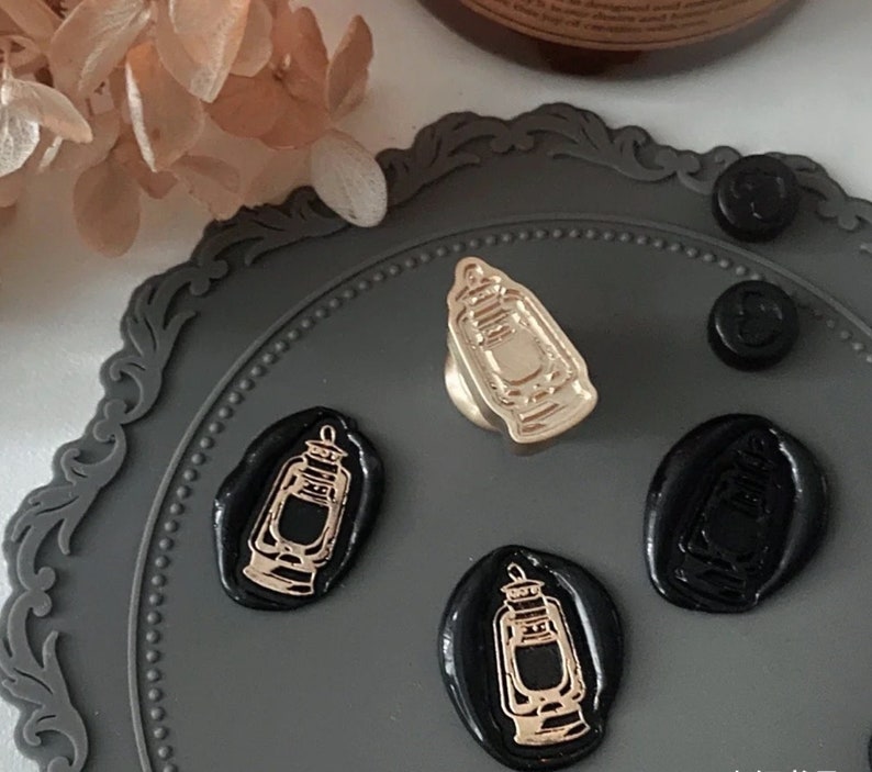 May include: A set of four black wax seals with a gold design of a lantern. The seals are on a gray surface with a decorative border.