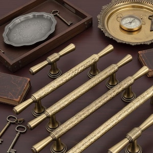May include: A selection of antique brass cabinet pulls with a hammered texture. The pulls are displayed alongside other vintage items, including a decorative tray, keys, and a small compass on a brass plate. The items are on a dark wood surface.