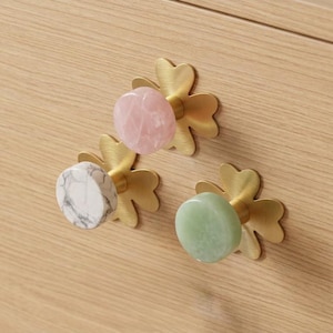 May include: Three decorative cabinet knobs with clover-shaped brass bases. The knobs feature polished stone in pink, white marble, and green. The knobs are mounted on a light wood cabinet.