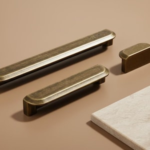 Könnte beinhalten: Satz antiker Messing-Schrankbeschläge, darunter zwei rechteckige Griffe und ein kleiner Knopf. Die Griffe sind unterschiedlich groß, mit abgerundeten Enden und einer leicht strukturierten Oberfläche. Die Beschläge werden vor neutralem Hintergrund gezeigt.