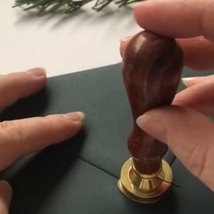 May include: A person is using a wooden handle wax seal stamp with a gold base to seal a dark green envelope.