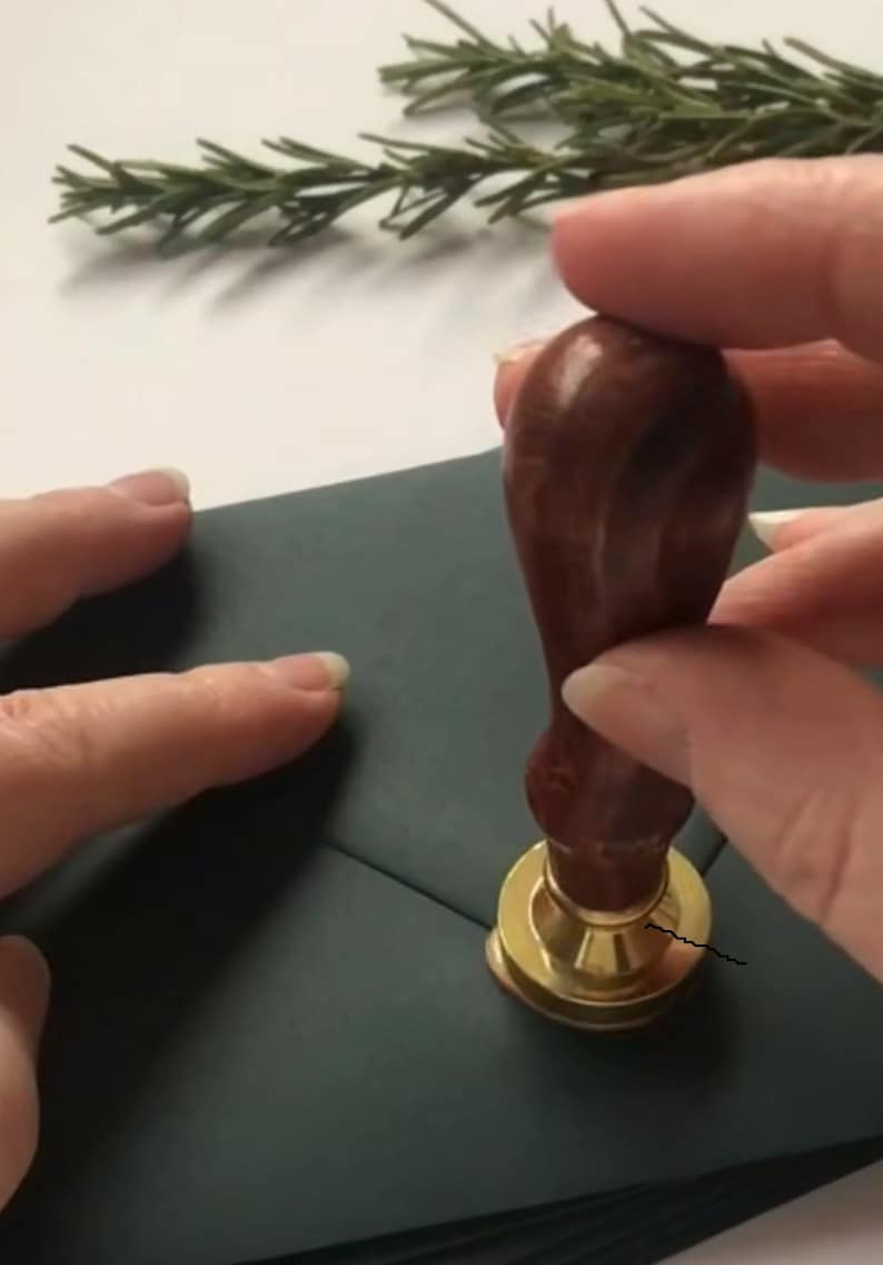May include: A person is using a wooden handle wax seal stamp with a gold base to seal a dark green envelope.