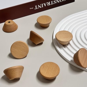 Könnte beinhalten: Eine Auswahl an Holzschrankknöpfen in verschiedenen Formen. Die Knöpfe sind aus hellem Holz gefertigt und weisen runde, konische und abgewinkelte Designs auf. Sie sind auf einer neutralen Oberfläche angeordnet, wodurch ihre natürliche Holzmaserung hervorgehoben wird.