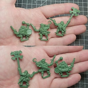 Puede incluir: Tres figuras en miniatura de plástico verde sin pintar que representan personajes barbudos con atuendo de pirata. Una figura sostiene un tambor y una maza, otra tiene una mano de gancho y una pistola, y la tercera lleva un gran bastón en forma de cruz.