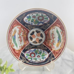 May include: A small, white ceramic bowl with a red, blue, and gold floral design. The bowl has a scalloped edge and is decorated with a variety of flowers, leaves, and birds.