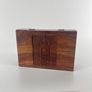 May include: A wooden box with a carved image of Big Ben on the front. The box is a dark brown color and has a hinged lid.