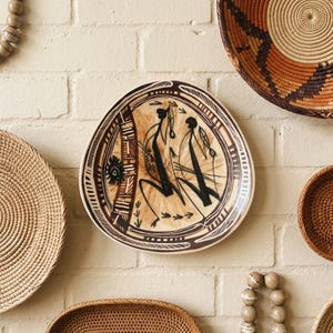 May include: A round ceramic decorative plate featuring a tan background with dark brown hand-painted abstract figures and geometric patterns. The design includes two stylized human-like silhouettes, vertical striped columns, and arrow motifs.