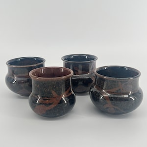 May include: Four small, handcrafted ceramic pots with a dark brown speckled glaze and a contrasting reddish-brown interior. The pots have a rounded shape and a wide opening, suitable for small plants or decorative use.