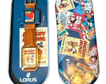 May include: A brown leather wristwatch with a gold-tone case and a Mickey Mouse design on the face. The watch is in a blue tin box with a Mickey Mouse design. The box has the words "Mickey Mouse" and "Lorus" printed on it.
