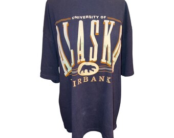 May include: A navy blue short-sleeve t-shirt featuring the text UNIVERSITY OF ALASKA FAIRBANKS in large white and yellow block letters. A graphic of a bear in a yellow oval is centered below the word ALASKA on the front of the garment.