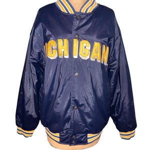 May include: A navy blue satin bomber jacket with the word "MICHIGAN" in yellow and white lettering across the chest. The jacket has a ribbed collar, cuffs, and hem with yellow and white stripes. The jacket is buttoned up.