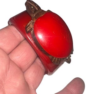 May include: A red bakelite bangle bracelet with a carved wooden turtle design. The turtle is on top of the bangle and has gold accents.