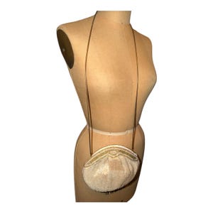 May include: A cream-colored, semi-circular handbag featuring intricate beadwork and sequins across the front surface. The bag includes a thin, metallic gold-toned shoulder strap attached to the top frame, which has a decorative gold-toned trim.