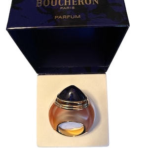May include: A Boucheron Paris Parfum bottle in a navy blue box. The perfume bottle is a ring shape with a dark blue top and a gold ring base. The box has the brand name and "PARFUM" printed on it.