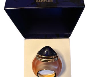 May include: A Boucheron Paris Parfum bottle in a navy blue box. The perfume bottle is a ring shape with a dark blue top and a gold ring base. The box has the brand name and "PARFUM" printed on it.