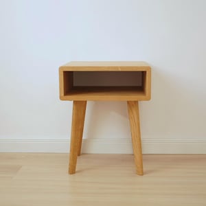 Peut inclure: Une table de chevet en bois clair dotée d'une section supérieure rectangulaire avec un compartiment de rangement ouvert et quatre pieds fuselés. Le meuble est fabriqué en bois naturel avec un grain visible et une finition lisse et claire.
