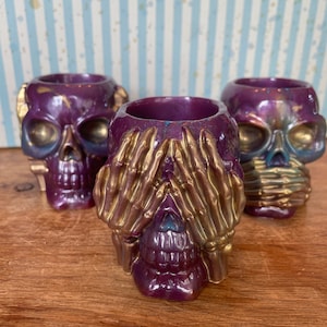 Puede incluir: Tres portavelas de calavera de color morado y dorado. La calavera del medio tiene manos que le cubren los ojos.