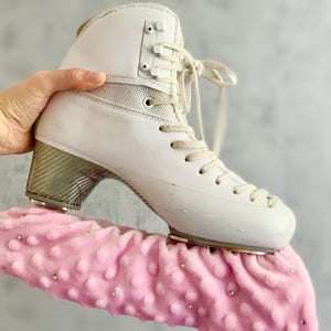 Op de afbeelding: Een witte schaats met een hak in metallic koolstofvezelstijl en witte veters, voorzien van een zachte, lichtroze textielhoes met textuur voor het ijzer, versierd met kleine, verspreide doorzichtige strass-steentjes die het ijzer omsluiten.