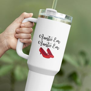 May include: White travel mug with a handle, clear lid, and straw. The mug features the text "Auntie Em Auntie Em" in black script, along with an illustration of red shoes. The mug is designed for cold beverages.
