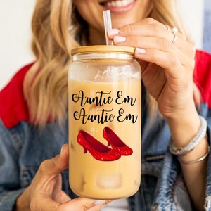 May include: A clear glass tumbler with a bamboo lid, filled with a light-colored beverage and ice cubes. The tumbler features the text "Auntie Em Auntie Em" in black script, along with a pair of red glitter high heels.