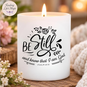 May include: A white cylindrical candle in a frosted glass container with a lit wick. The front features black text that reads "BE Still and know that I am God" with "PSALM 46:10" below it, accented by a small floral graphic design above the text.