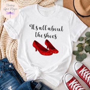 May include: A white short-sleeved t-shirt featuring the black text It's all about the shoes printed above a graphic of two red glittery high-heeled pumps. The shirt has a crew neckline and a casual fit, showcasing a simple and clean design.