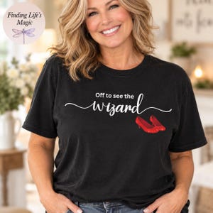 May include: A black short-sleeved t-shirt featuring the white text "Off to see the wizard" in a cursive script font. Below the text, there is a graphic illustration of a pair of shiny red high-heeled shoes positioned side by side.