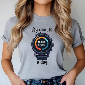 May include: A light gray t-shirt featuring a graphic of a black digital watch face with a circular progress ring in orange and teal. The text above the watch reads My goal is and the text below reads a day, with 10,000 STEPS in the center.