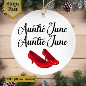 May include: White ceramic ornament with the text "Auntie June Auntie June" in black script. Two red high-heeled shoes are at the bottom. A gold string is attached for hanging. The ornament is set against a background of pine needles and wood.