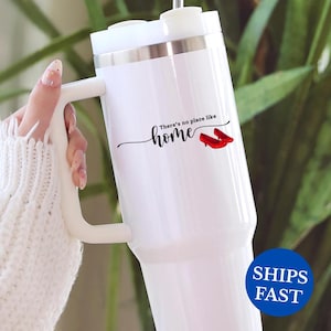 May include: White travel mug with a handle and a silver band near the top. The mug features the text "There's no place like home" with an illustration of red shoes. The mug has a white lid.