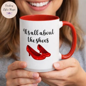May include: A white ceramic mug with a red interior and handle, featuring the black text It's all about the shoes and an illustration of two sparkling red high-heeled pumps on the front. The design is centered on the exterior of the mug.