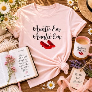 May include: A light pink t-shirt with the text "Auntie Em Auntie Em" and a pair of red glitter shoes. A matching mug and a tag that says "For the best Auntie!" are also visible. A book with the text "She is clothed with strength and dignity" is open.