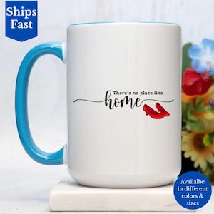 May include: White ceramic mug with a blue handle and rim. The mug features the text "There's no place like home" with an illustration of red shoes. The mug is available in different colors and sizes.