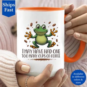 May include: White ceramic coffee mug with an orange interior and handle. The mug features a cartoon frog illustration and the text "I MAY HAVE HAD ONE TOO MANY CUPS OF COFFEE". The mug is held by a person.