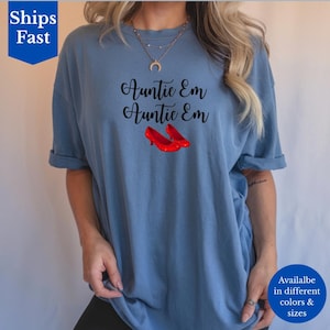 May include: Auntie Em graphic tee in a dusty blue color. The shirt features the text "Auntie Em Auntie Em" in black script, with a pair of red high-heeled shoes below the text. The shirt is available in different colors and sizes.