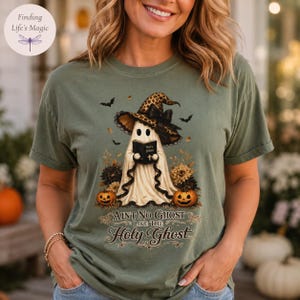 May include: An olive green t-shirt featuring a graphic of a ghost wearing a leopard print witch hat and holding a book labeled Holy Bible. The design includes pumpkins, flowers, bats, and the text AINT NO GHOST LIKE THE HOLY GHOST.