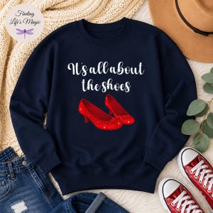 May include: A navy blue crewneck sweatshirt featuring the white text It's all about the shoes centered above a graphic illustration of a pair of sparkling red high-heeled pumps. The design is printed on the front of the long-sleeved pullover garment.
