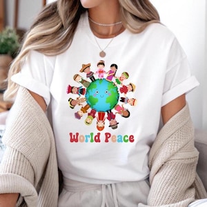 May include: White t-shirt featuring a colorful graphic of children from around the world surrounding a smiling globe. The words "World Peace" are printed in rainbow colors below the globe. The shirt has short sleeves.