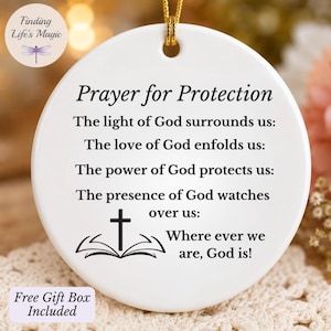 May include: A round white ceramic ornament featuring black printed text that reads Prayer for Protection followed by a religious verse and a graphic of a cross above an open book. A gold string is attached to the top for hanging purposes.