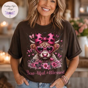 May include: A dark brown t-shirt featuring a graphic of a Highland cow wearing pink heart-shaped sunglasses and a large pink cow-print bow. The design includes pink butterflies, flowers, and the text "Bow-tiful & Blessed" in pink script lettering.